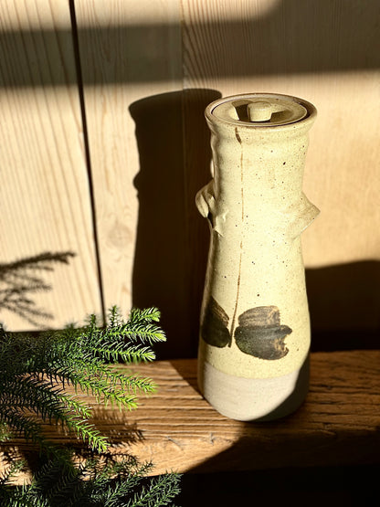 A tall hand-thrown ceramic pot with a matt buff colorued glaze and hand-painted design. It has visible handles and is placed next to some greenery.
